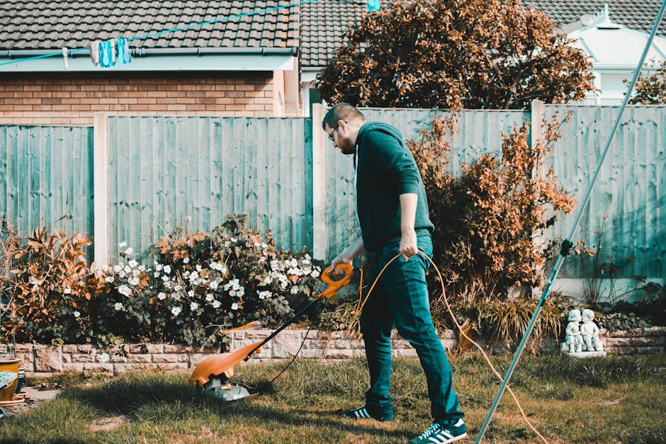 A man wearing glasses, a dark green hoodie, and blue jeans is trimming grass in a residential garden with an electric grass trimmer, standing on a lush green lawn. The garden features a well-maintaine